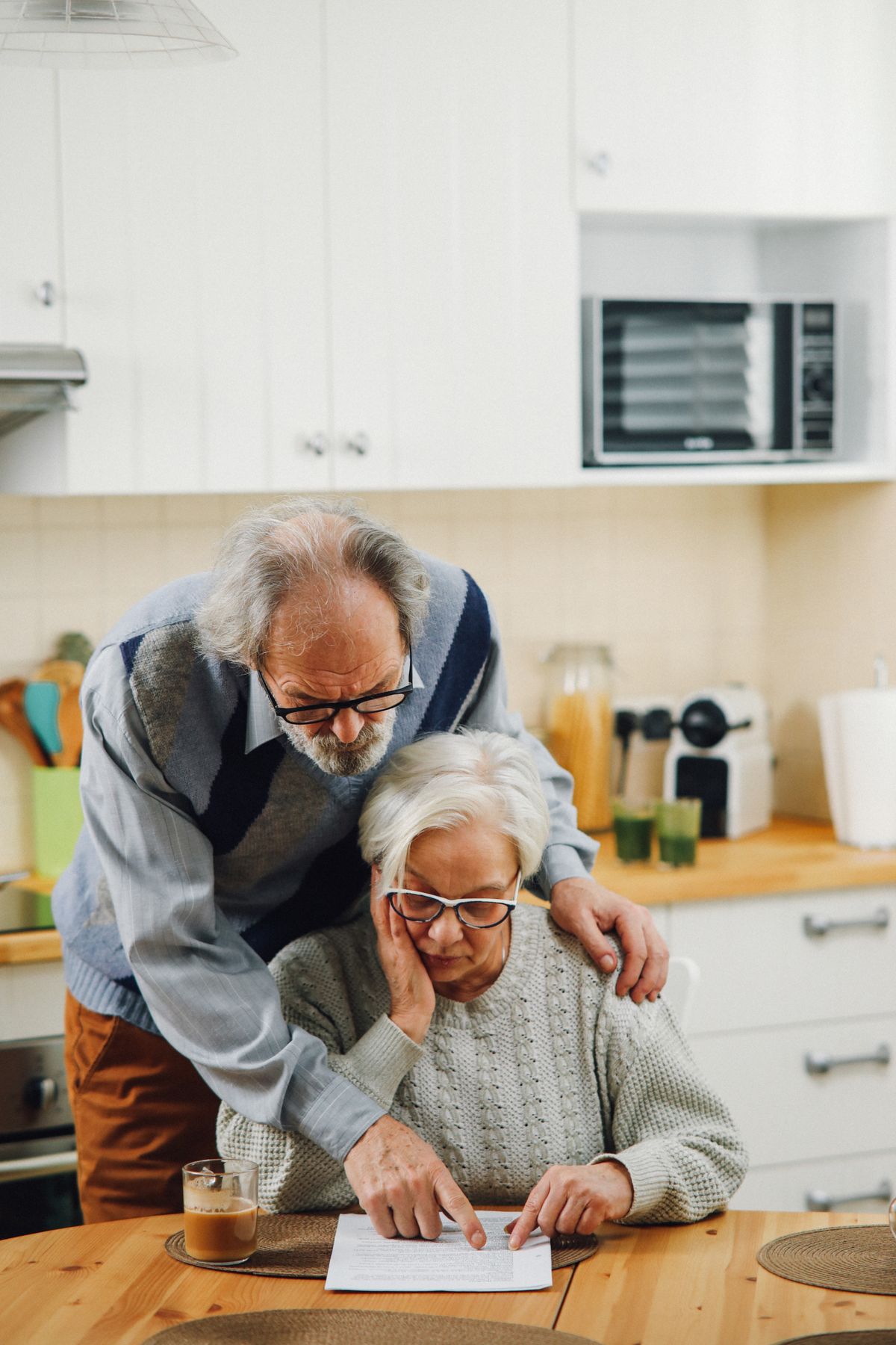 Two people sitting together at a kitchen table, one helping the other with paperwork, warm natural light through a window — representing informal family support in a home setting