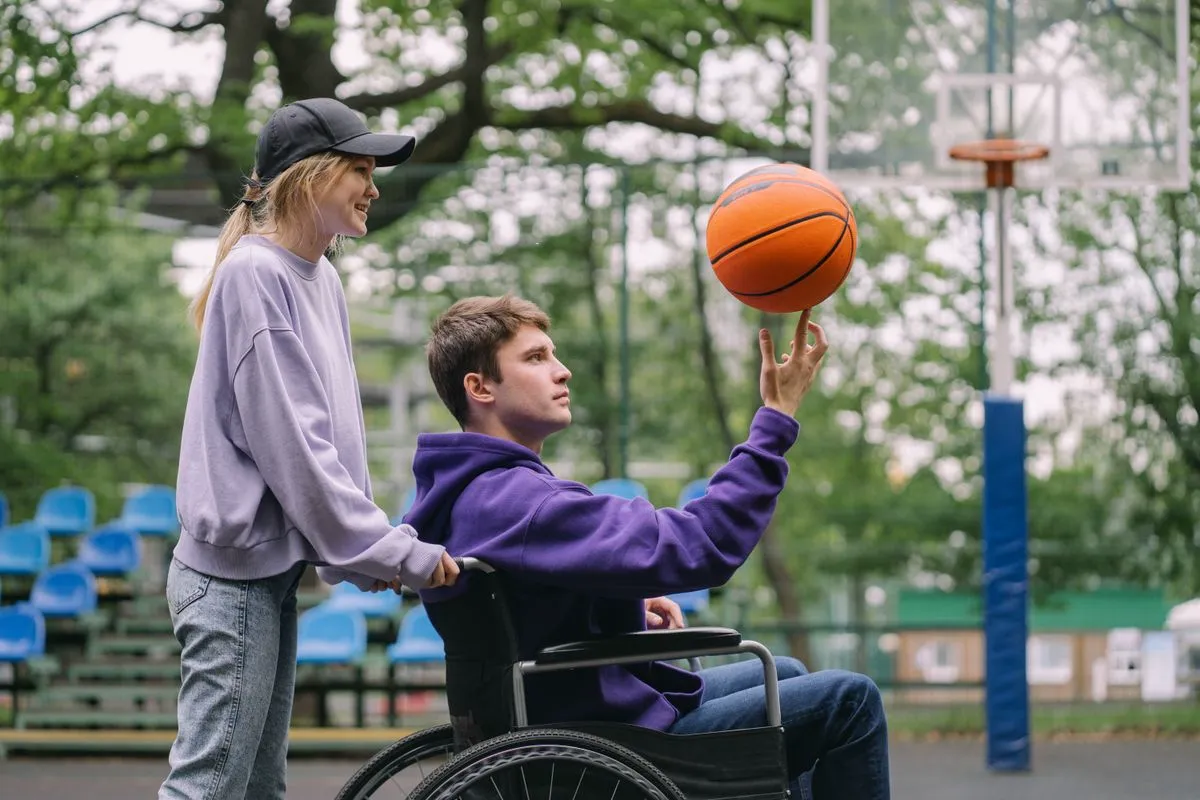 Young man in wheelchair spinning a basketball while a supportive friend watches, active community participation